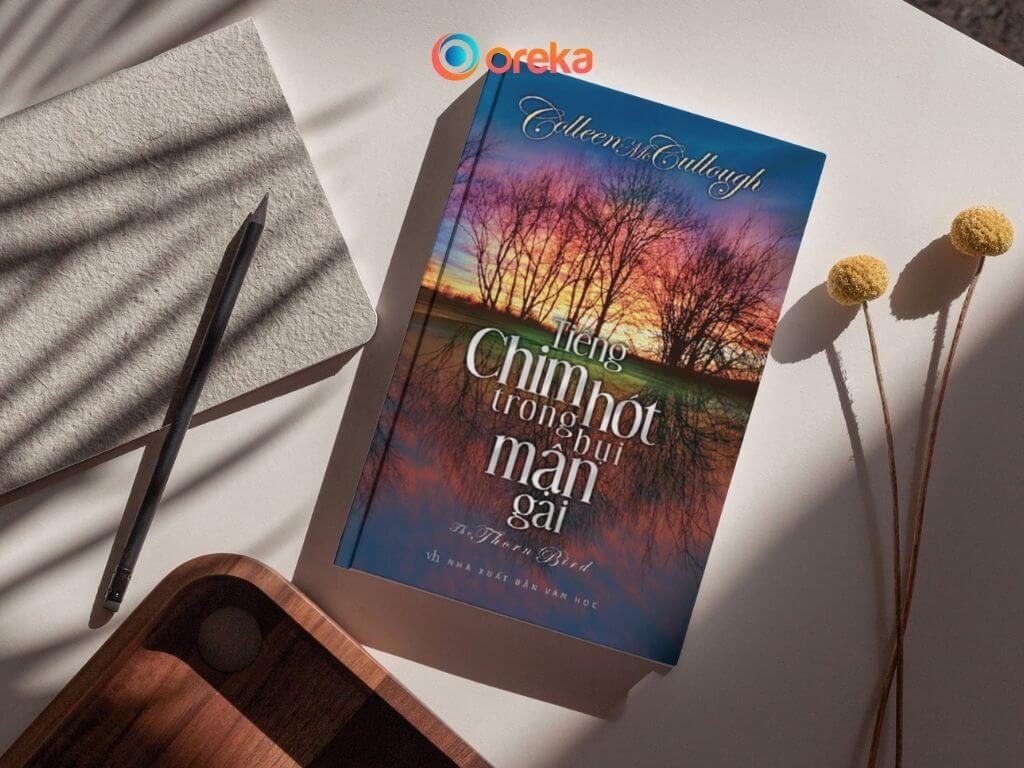 review Tiếng chim hót trong bụi mận gai, hình ảnh bìa sách được đặt trên mặt bàn, bên cạnh có 1 cuốn sổ, 1 cái bút, 1/4 khay gỗ và hai cành hoa khô