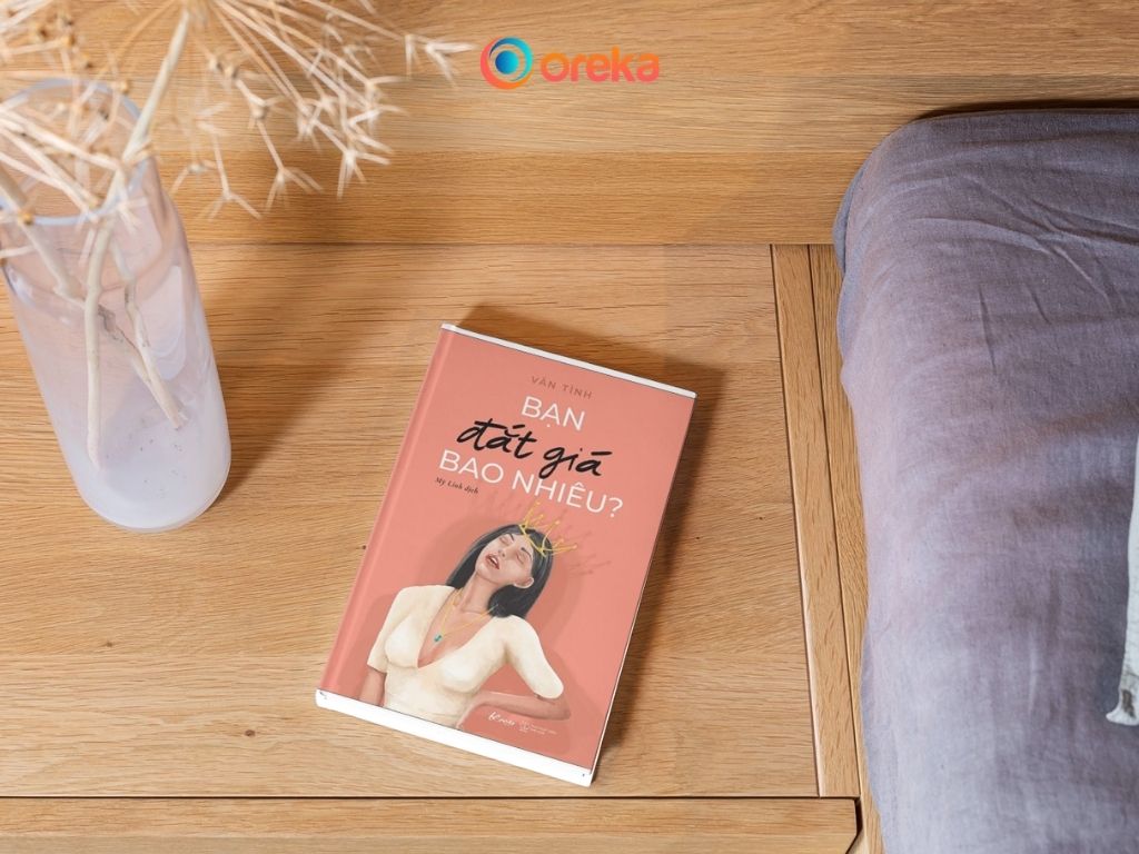 Bạn đắt giá bao nhiêu review, hình ảnh cuốn sách được đặt tại tủ gỗ đầu giường bên cạnh có một bình hoa khô.